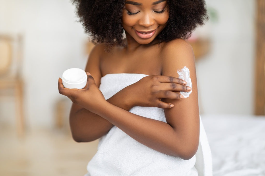 Woman at home applying cream and nourishing body butter while wearing towel after bath.