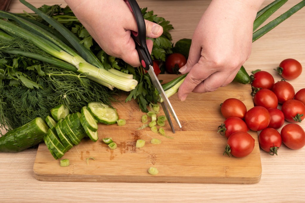 Woman cuts green onion with scissors over a cutting board. Greens, tomatoes and cucumbers on a wooden table. Rustic style