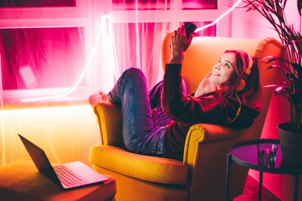 a-woman-smiling-holding-her-phone-sitting-on-a-sofa-a-laptop-on-a-table-and-LED-lights-with-different-colors.