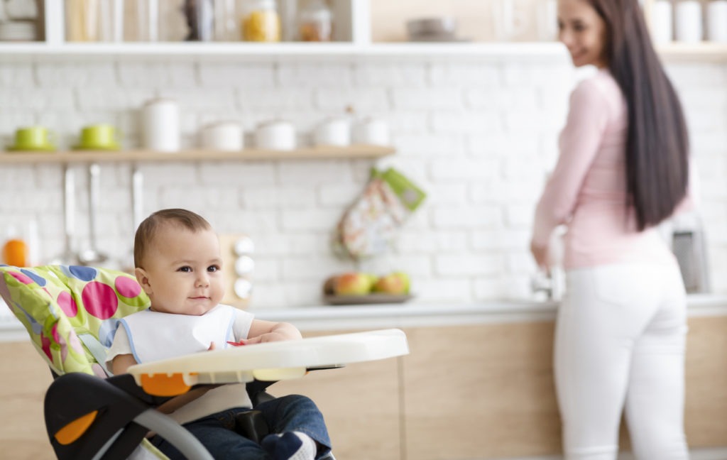 Cute baby playing on high chair in kitchen, mother washing dishes