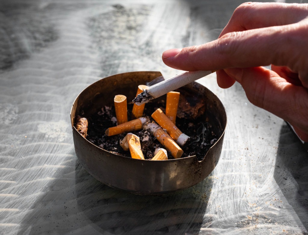 Hand holding a cigarette whose ashes are tapped into an ashtray