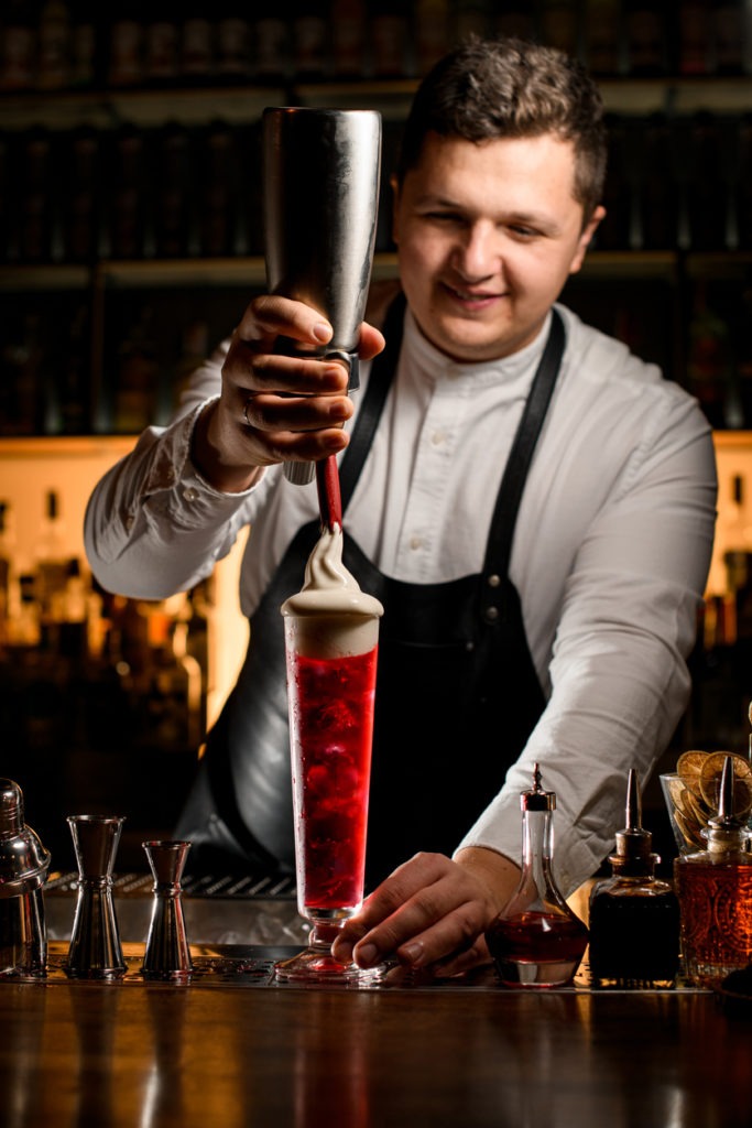 male bartender neatly decorates a tall transparent glass of bright alcoholic cocktail with whipped cream
