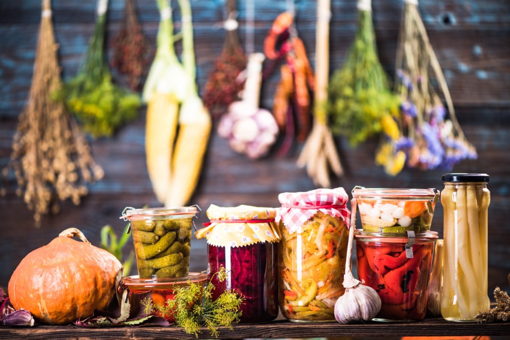 Different sizes of canning jars containing different types of fermented food on the table with vegetables hanging on the wall in the background
