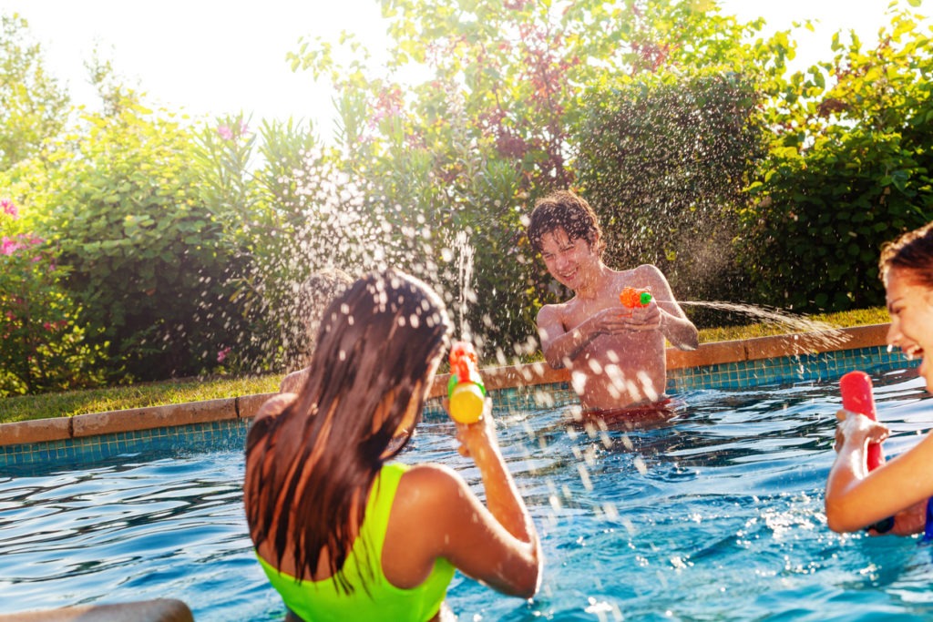 Group of three teens plays with water-gun squirt pistol on swimming pool outside on a sunny day