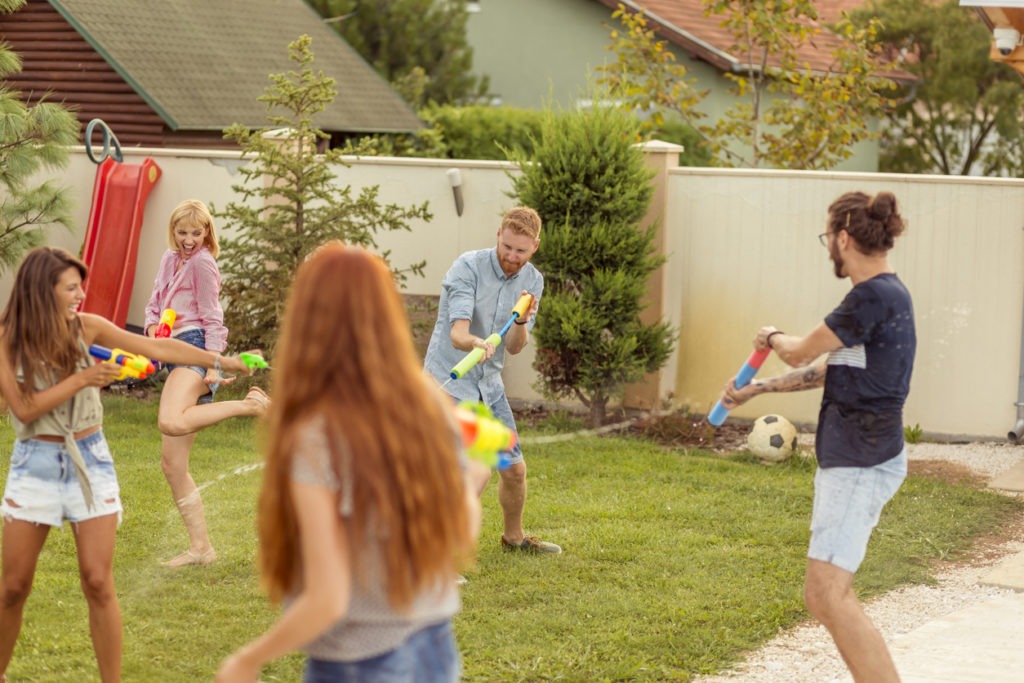 Group of young friends having fun spending summer day outdoors, playing with squirt guns, splashing water on each other, running, and chasing each other