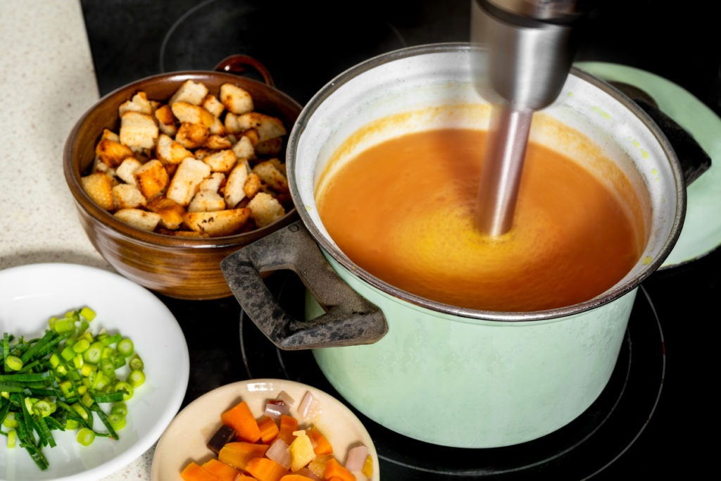 Hand-blender-in-a-pot-on-stove-mixes-carrot-soup-plates-with-vegetable-and-sliced-fried-bread-on-kitchen-board.-Preparation-of-fresh-carrot-soup