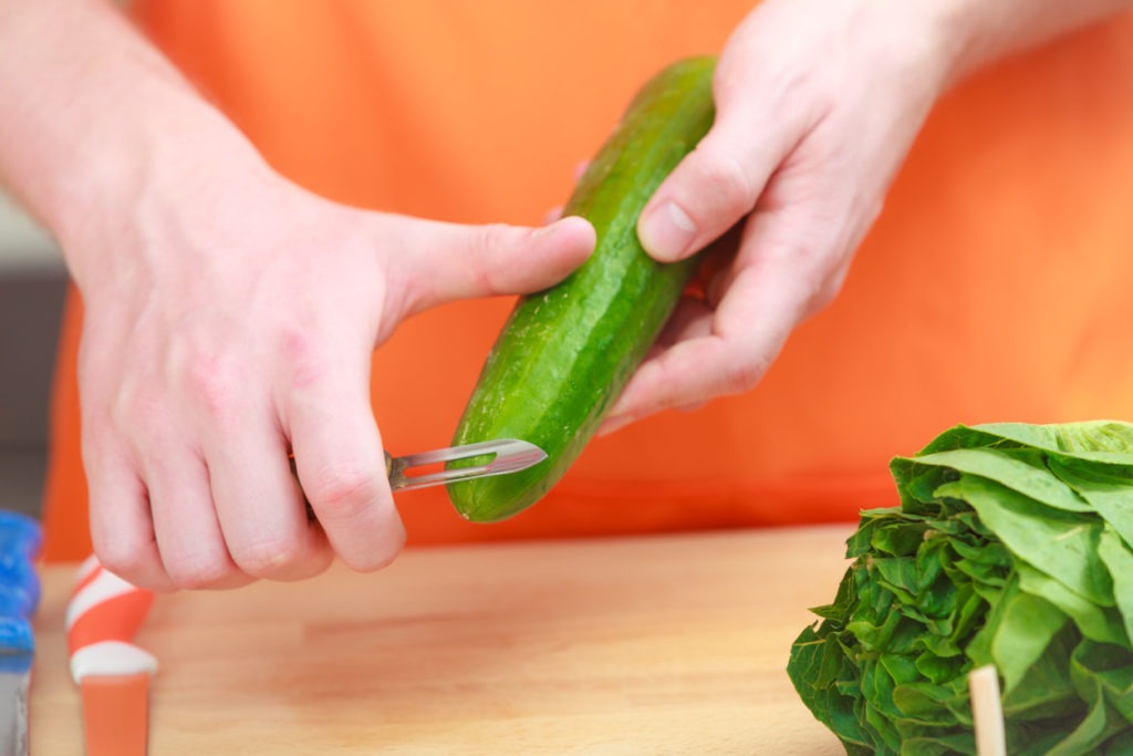 Man wearing an orange apron is preparing vegetable salad by peeling cucumber with a swivel peeler