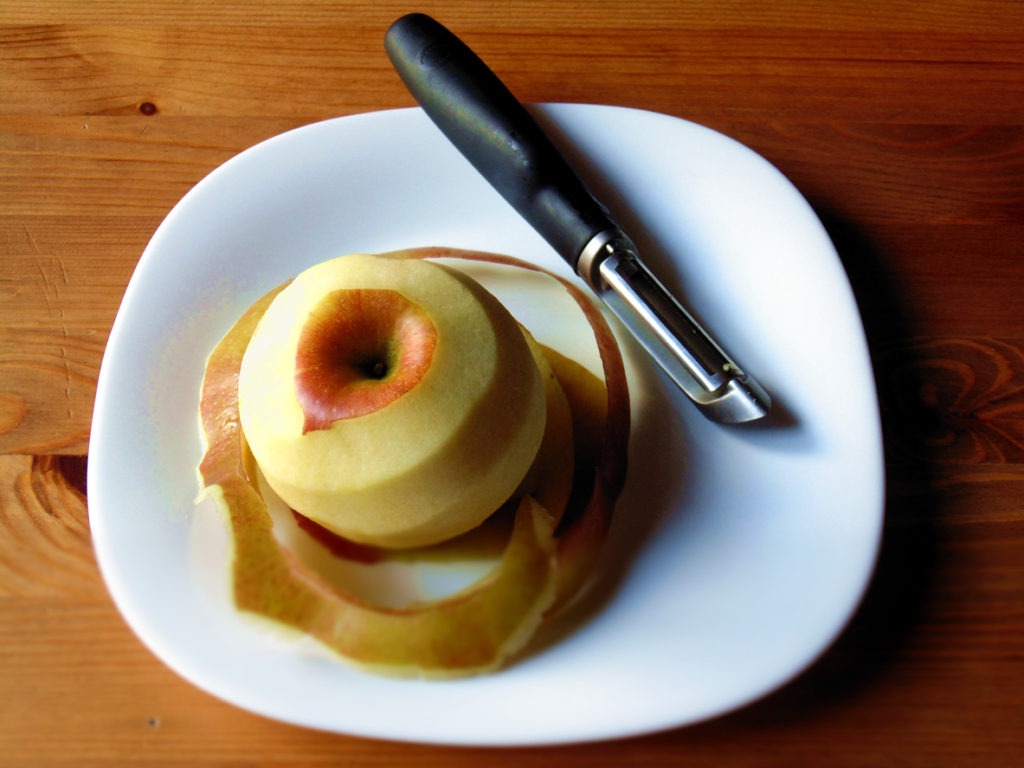 Peeled red apple beside a swivel peeler on a white plate on a wooden table