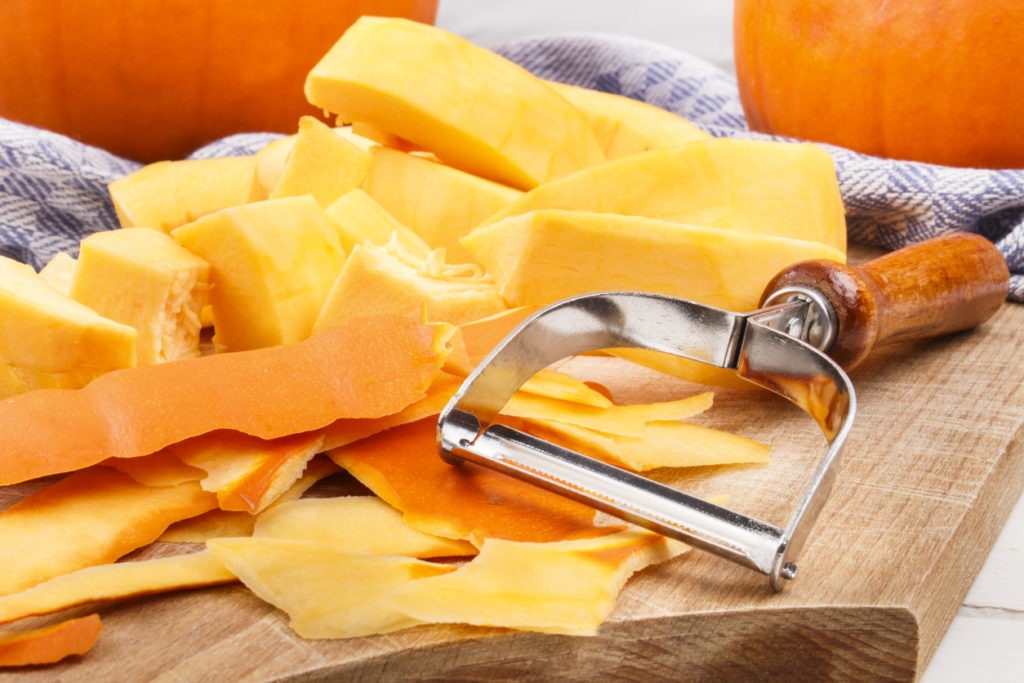 Slices of peeled pumpkins beside pumpkin peelings and a serrated peeler on top of a chopping board