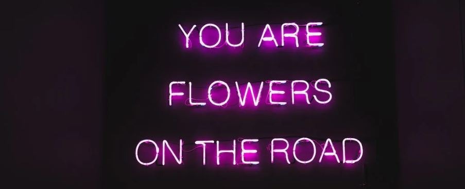 a neon pink sign on a wall with “you are flowers on the road” written