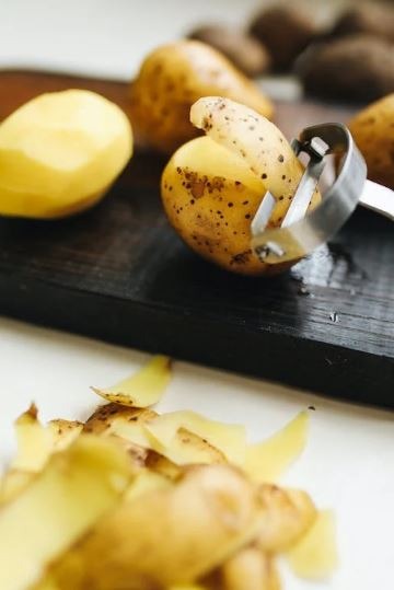a potato being peeled with a Julienne peeler beside a peeled potato and its shavings