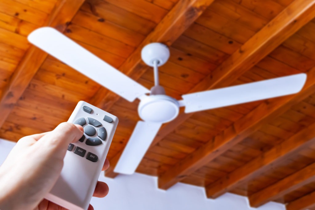 Close up shot of a hand using a remote control to operate a ceiling fan mounted in a house on a wooden ceiling