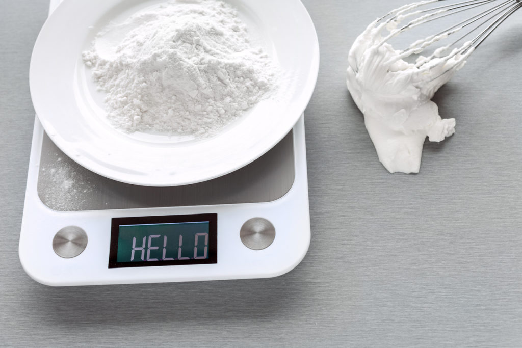 a whisk with batter beside a kitchen scale with a plate of flour on top and an inscription on the scale displaying: "hello"