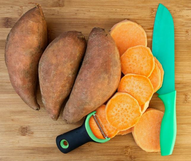 a whole sweet potato and sliced sweet potatoes beside a knife and a y-shaped peeler