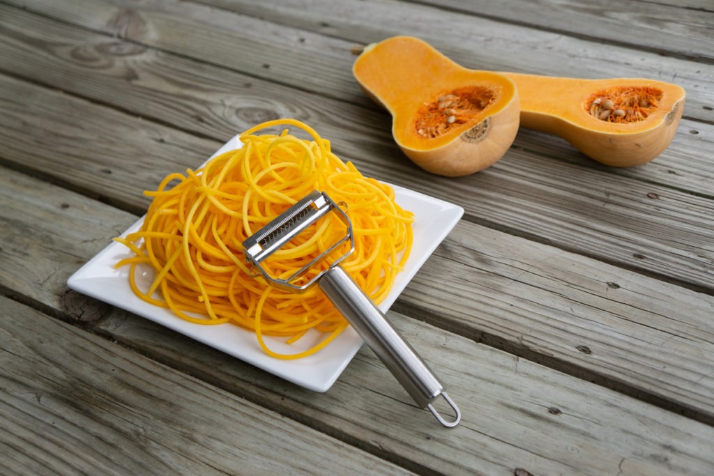 freshly peeled butternut squash vegetable noodles on a plate with a Julienne peeler beside a sliced butternut squash