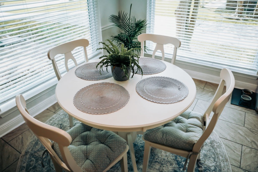 Close-up of a small table and four chairs in an eat-in kitchen with a tile floor, a rug, a plant, and placemats