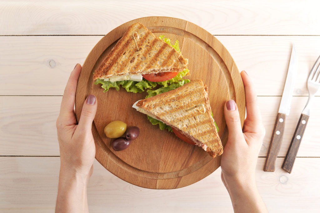 Club sandwich of fried white bread with a tuna filling on a wooden tray. The sandwich is cut into two halves. The tray is held in hand. There is cutlery nearby. View from above. Close-up