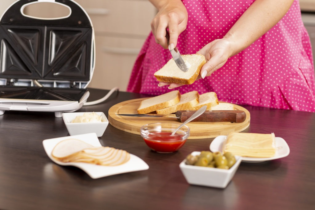 Detail of female hands spreading butter over a bread slice; woman making hot sandwiches in a sandwich maker for breakfast. Focus on the knife