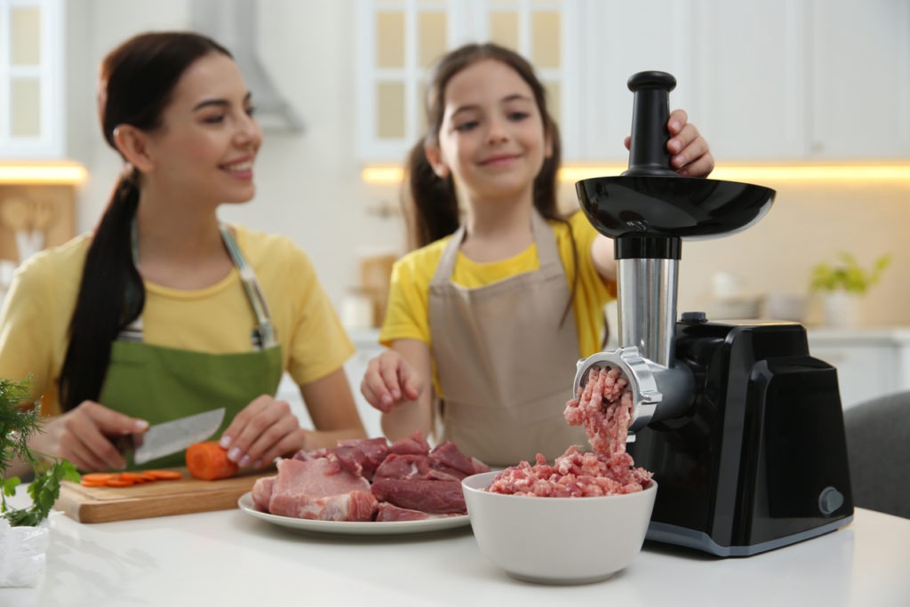 Happy family making dinner together in kitchen, daughter using modern meat grinder while mother cutting carrot