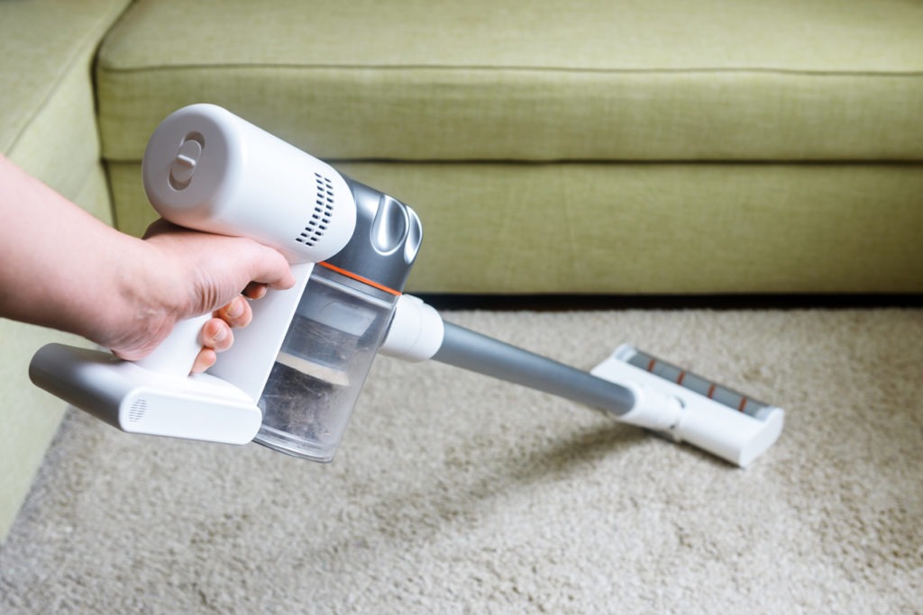 Wireless vacuum cleaner used on carpet in room. Housework with new white hoover. Person holds modern vacuum cleaner by sofa