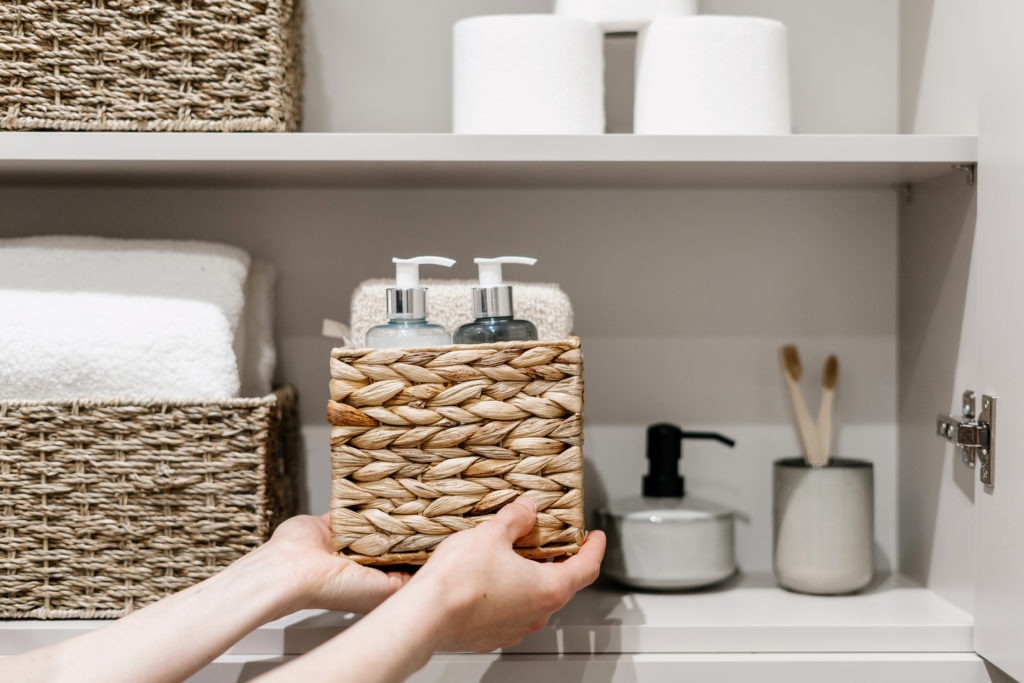 woman-putting-wicker-box-with-bath-sponge-shampoo-soap-dispenser-bottle-and-other-cosmetics-products-in-closet