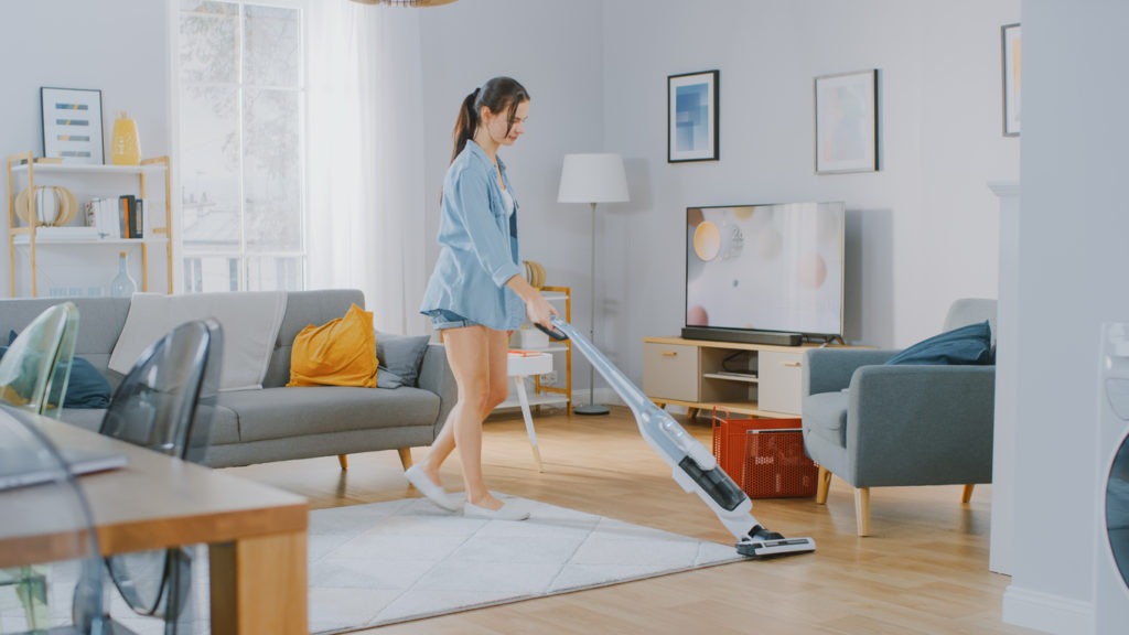 Young Beautiful Woman in Jeans Shirt and Shorts is Vacuum Cleaning a Carpet in a Bright Cozy Room at Home. She Uses a Modern Cordless Vacuum. She's Happy and Cheerful