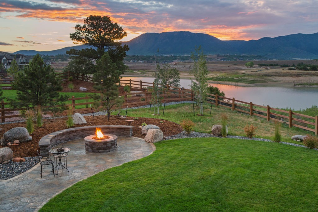 A backyard with a stone fire pit and a scenic view of a river and some mountains during sunset.
