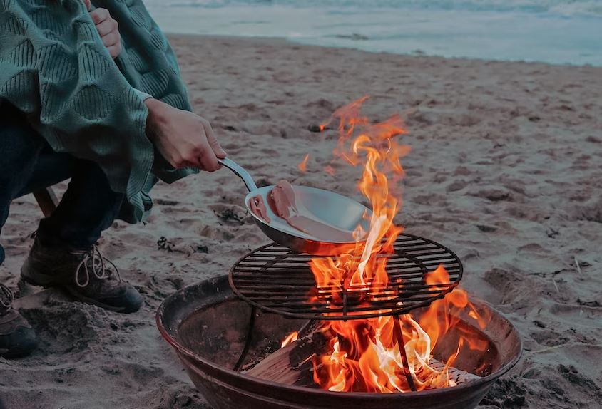 A person cooking meat over a portable fire pit that has a grill on top.