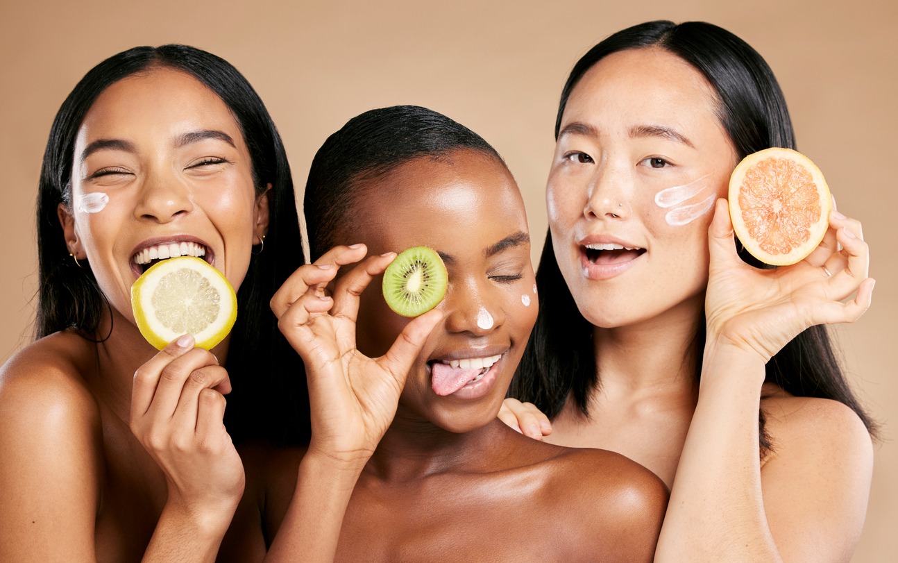 a close-up image of three multiracial women, with dabs of cream on their faces, goofily holding fruits
