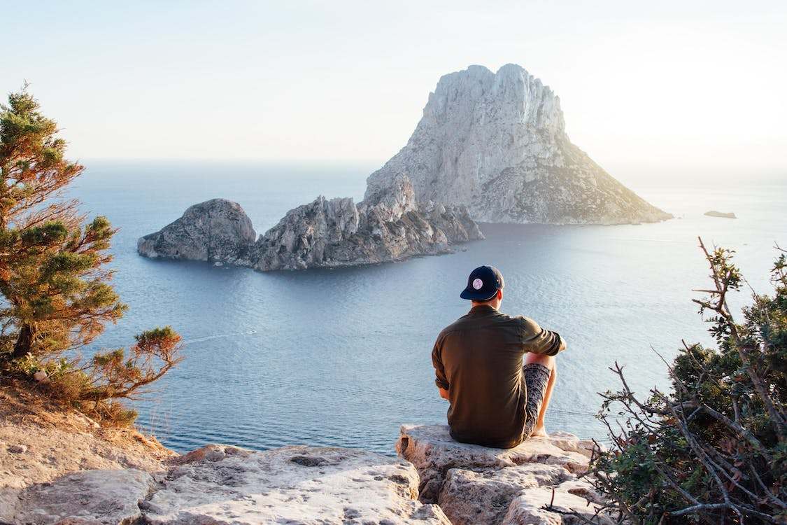 A man wearing a cap sitting on an edge facing the sea