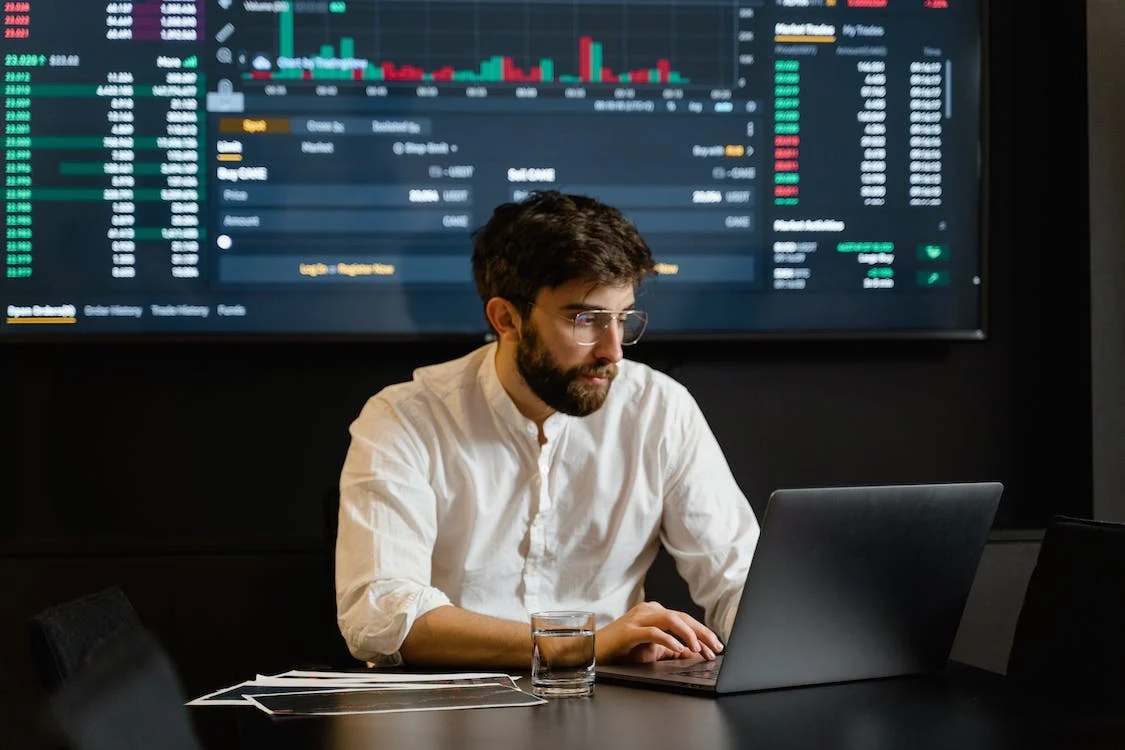 A man wearing white polo using laptop, with trading graphs as his background
