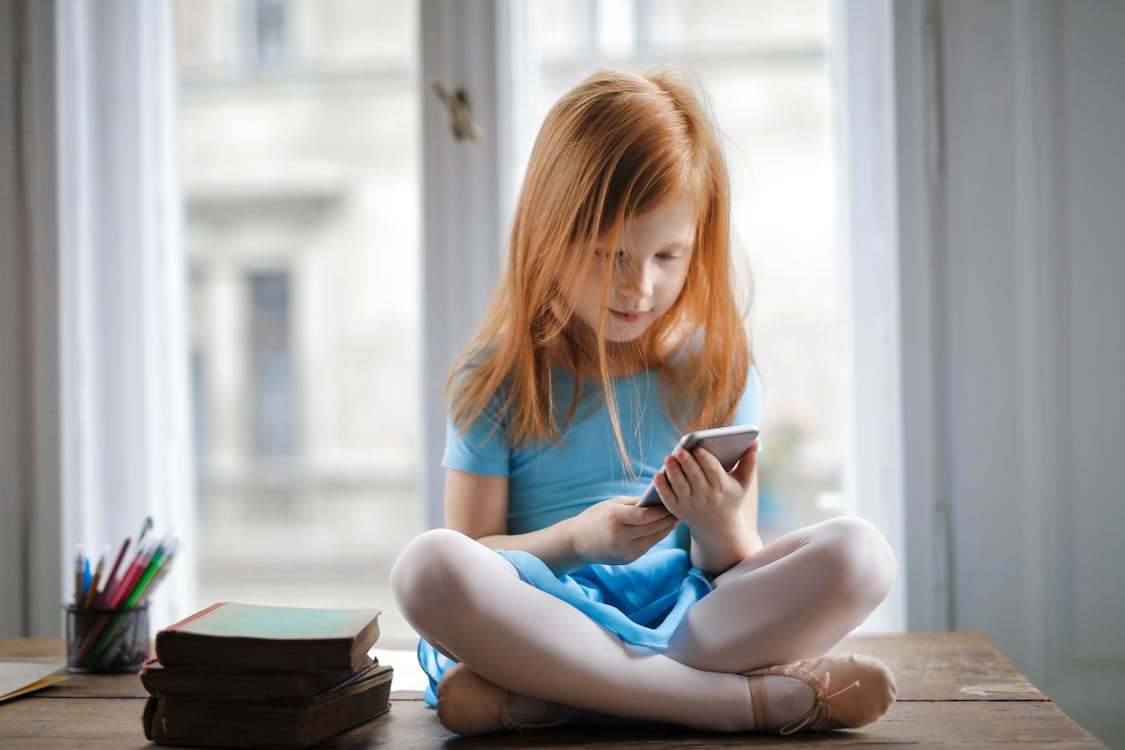 A photo of a small girl sitting on table and using smartphone in light living room