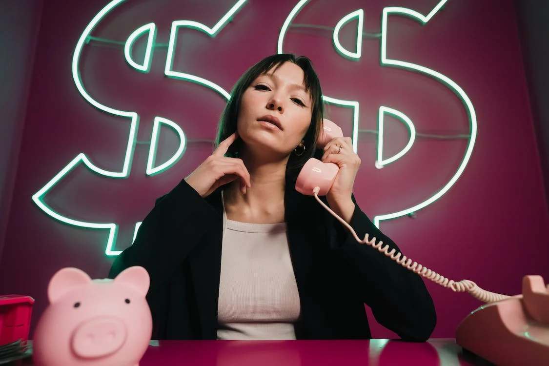 A woman holding a pink telephone, with massive dollar signs as her background