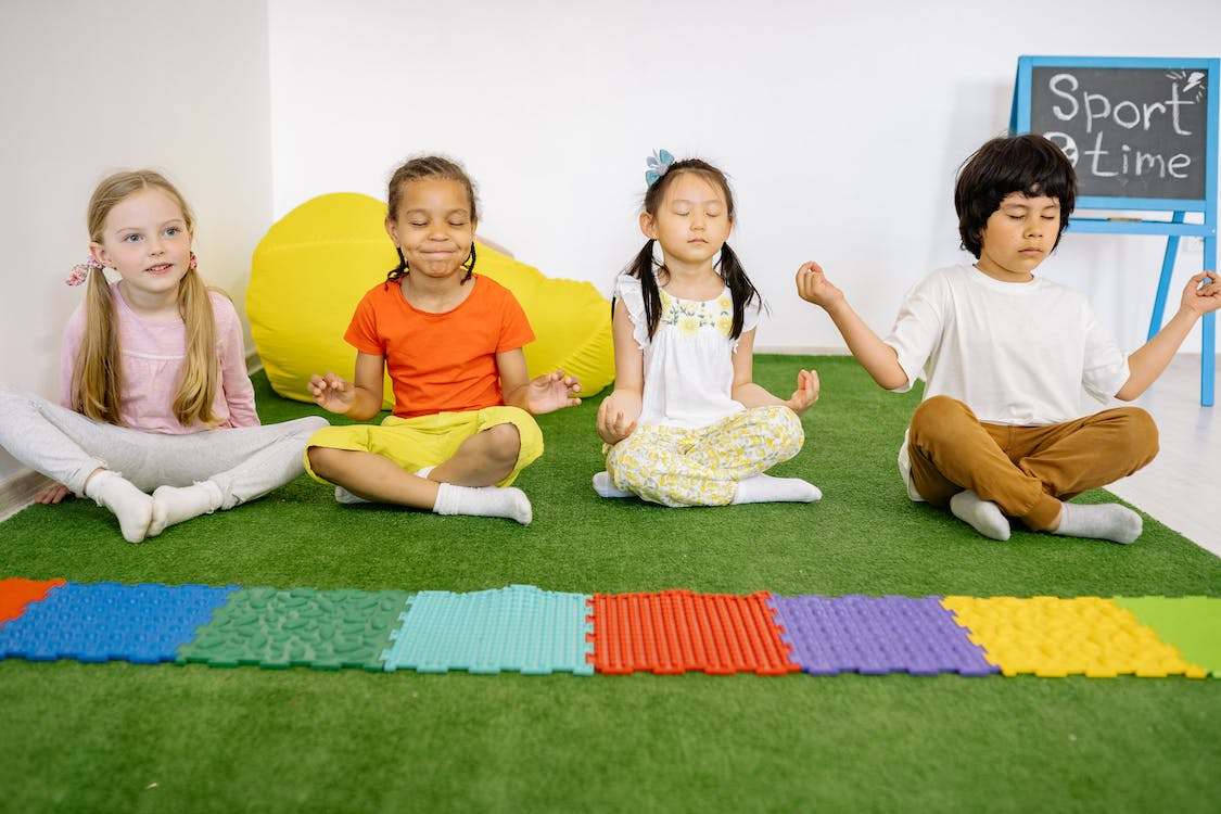 Four children sitting on a green mat while doing yoga poses