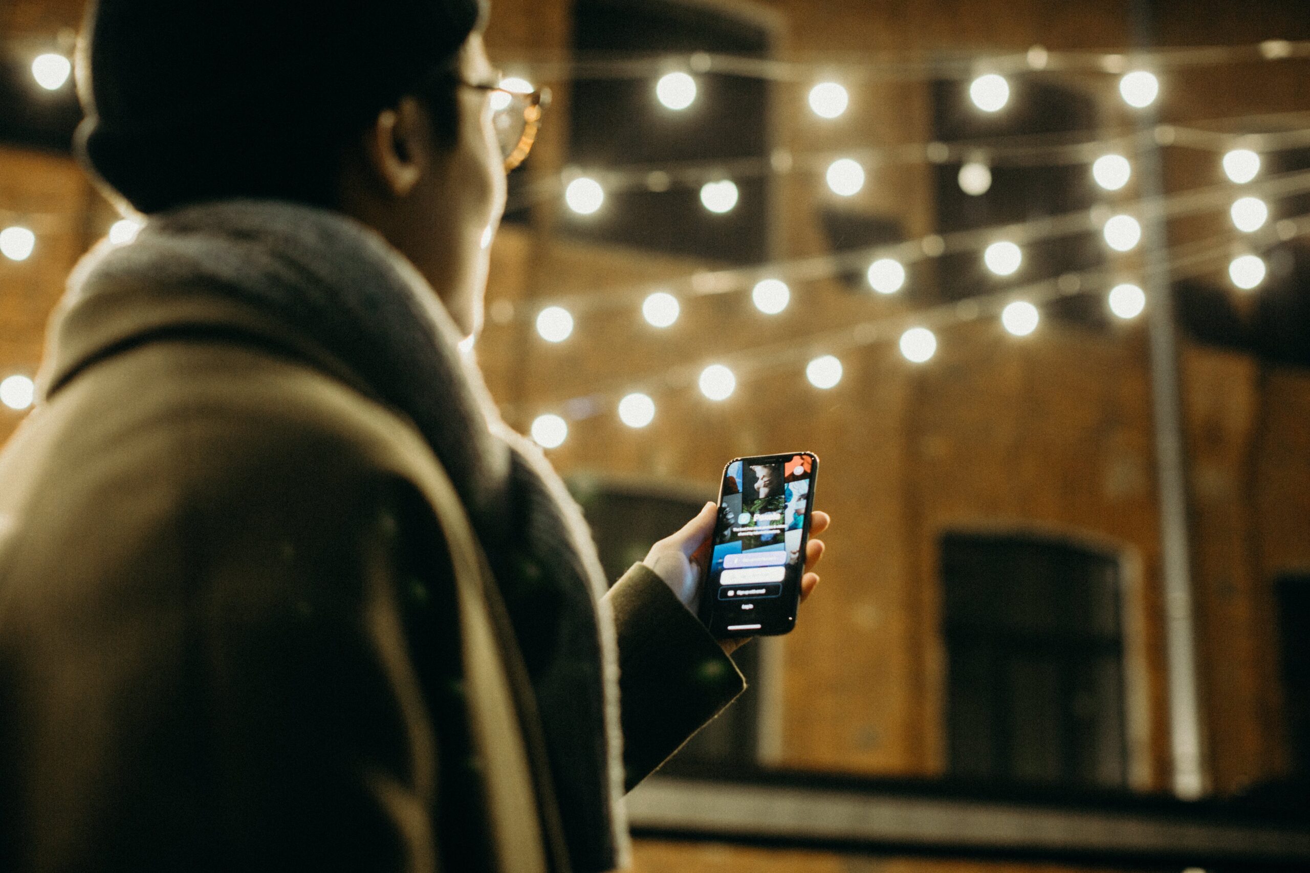 selective-focus-photography-of-man-using-smartphone-beside-string-lights