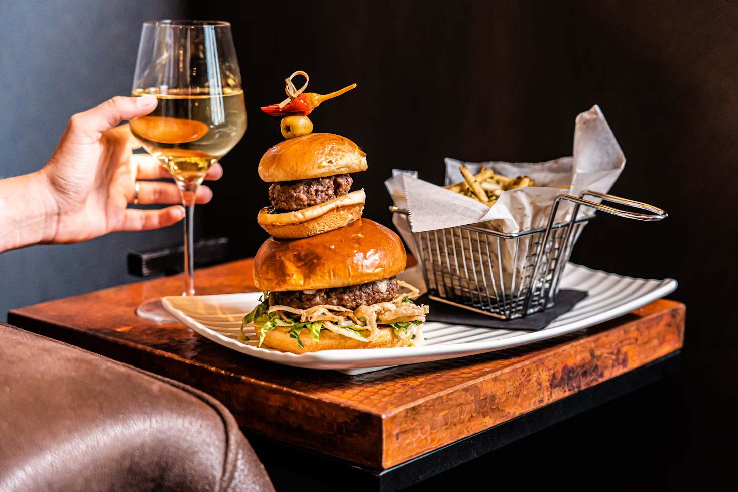Two-tier burgers, fries and a glass of wine on a table with a person’s hand
